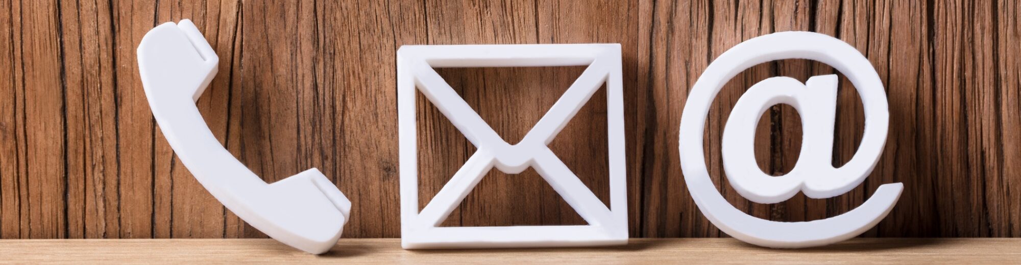 Drei weiße 3D-Symbole für Telefon, E-Mail und Kontakt (Hörer, Briefumschlag, At-Zeichen) vor einem Hintergrund aus Holzlatten